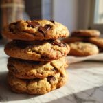 A stack of four freshly baked chocolate chip cookies, with more cookies blurred in the background.