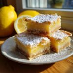 Three lemon bars 9x13 pan stacked on a plate, dusted with powdered sugar, with lemons in the background.