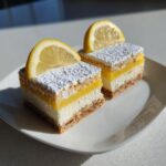 Two lemon squares topped with powdered sugar and a lemon slice, sitting on a white plate.