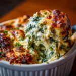 Close-up of creamy spinach artichoke dip being scooped from a white dish, showing melted cheese and spinach.