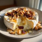 Baked brie cheese topped with nuts, dried fruit, and honey, oozing from a cut wedge.