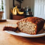 A loaf of banana bread with a slice cut, sitting on a white plate. Ripe bananas are in the background.