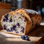 A loaf of blueberry lemon sourdough bread sits on a wooden board with scattered blueberries.