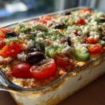 A vibrant Greek Layer Dip in a glass baking dish, topped with tomatoes, cucumbers, olives, feta, and dill.