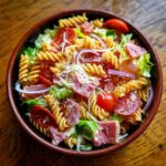 Overhead shot of a colorful grinder pasta salad with rotini, salami, lettuce, tomatoes, and red onion.