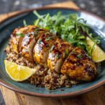 A plate of honey mustard chicken sliced and served over rice with a side salad and lemon wedges.