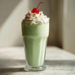 A vibrant green shamrock shake in a glass, topped with whipped cream and a cherry.