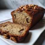 A loaf of vegan banana bread, a perfect addition to bridal shower desserts, with a slice cut and walnuts on top.