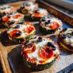 Close-up of several Zucchini Pizza Bites on a baking sheet, topped with pepperoni, cheese, and olives.