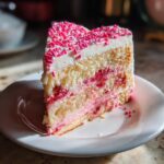 A slice of heart-shaped gluten-free Valentine desserts cake with pink sprinkles, frosting, and raspberry filling on a white plate.