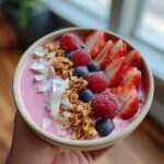 A vibrant healthy pink smoothie bowl topped with fresh strawberries, raspberries, blueberries, granola, and coconut flakes.
