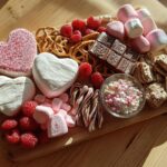 A Valentine’s S’mores board featuring heart-shaped marshmallows, chocolate, pretzels, raspberries, and candy.